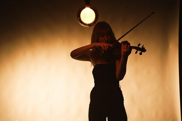 Person playing the violin under a spotlight with a warm, illuminated background, identified as performer Emma Greenhill.