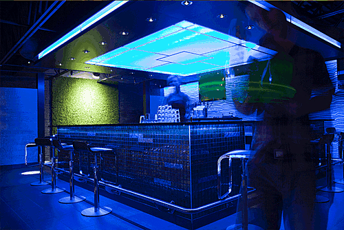 Modern bar with blue lighting, tiled counter, stools and two people slightly blurred in motion.