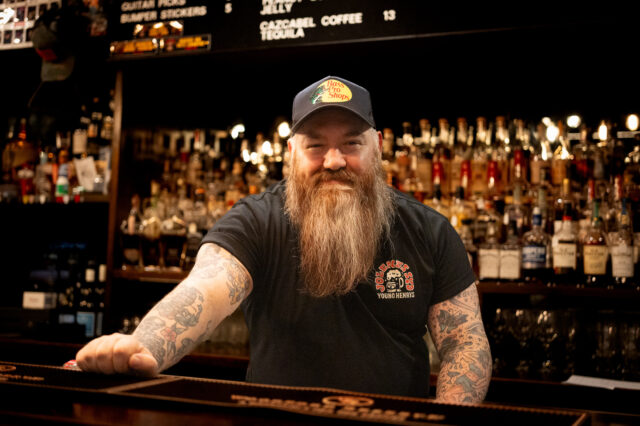 Bearded person with tattooed arms behind a bar counter, wearing a 'YOUNG HENRYS' t-shirt and cap; shelves with bottles and a menu board in the background.