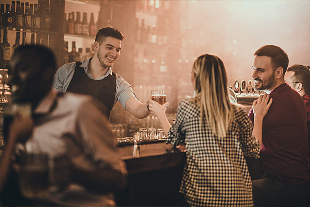 Bartender handing a drink to a customer in a busy bar with shelves of bottles in the background.