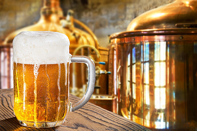 Glass mug of beer with frothy head on a wooden surface in front of copper brewing tanks.