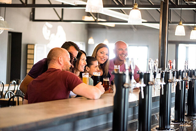 Group of people sitting at a bar counter with beer taps in the foreground and hanging lights in the background.