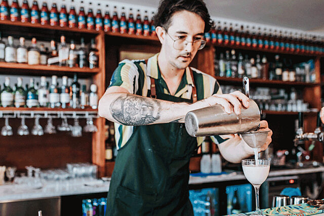 Bartender at Rosella's Bar pouring a drink from a cocktail shaker with shelves of alcohol and hanging wine glasses in the background.