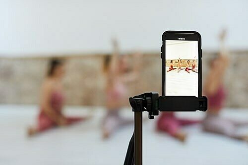 Smartphone on a tripod recording a yoga or exercise session with three blurred participants in the background, visible clearly on the phone screen.