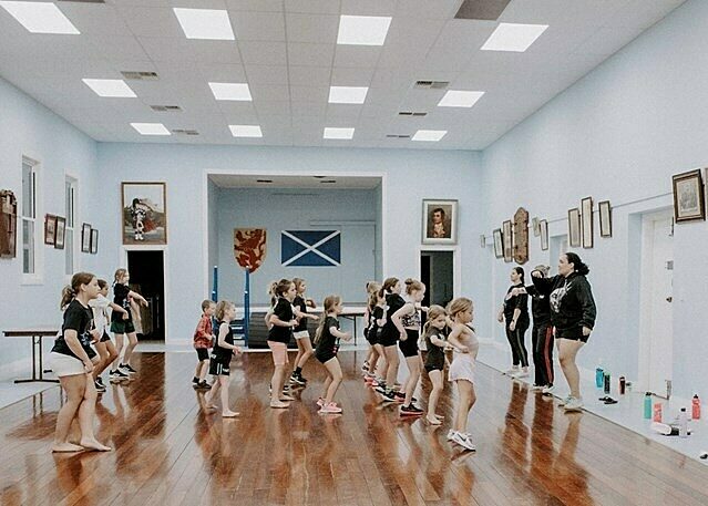 Group of children and adults participating in a dance or exercise class in a studio with wooden floors, light blue walls, and a Scottish flag.