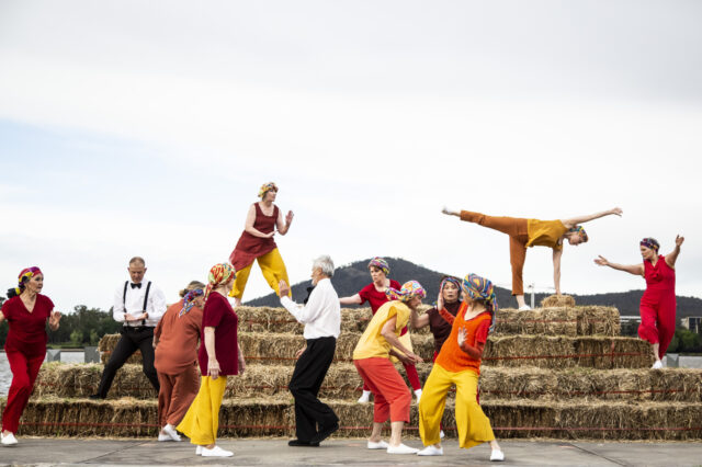 Group of dancers from Canberra Dance Theatre performing in colourful outfits on a structure made of hay bales.