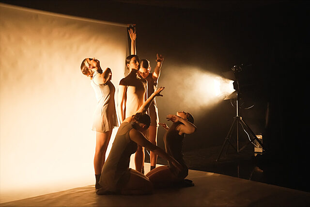 Made By performance still 2023. Group of people posing with raised arms in a dimly lit room, backlit by a bright light casting dramatic shadows.