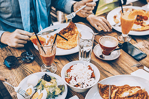 Table set with plates of salads, omelets or crepes, and a bowl of yoghurt with fruit and granola, surrounded by people enjoying coffee, juice, and water.