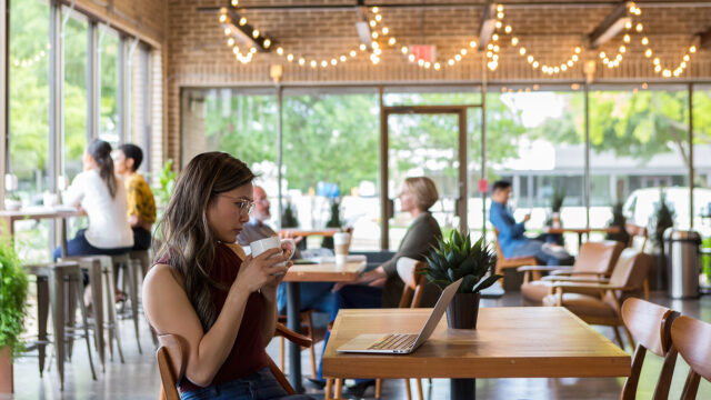 Person using a laptop and drinking from a cup in a sunlit cafe with others working or chatting at nearby tables.