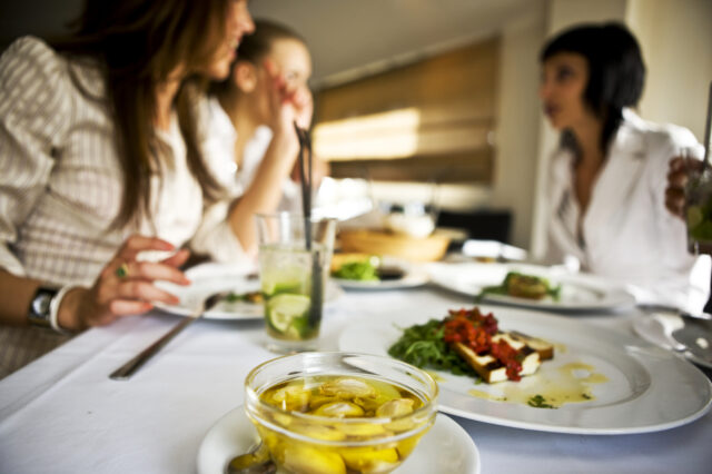Two people seated at a table with plates of food and drinks, including greens with tomato topping and yellow items in liquid.