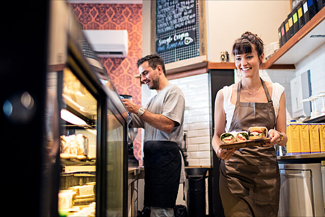 Two people in a café or restaurant, one holding a tray of food and the other near a counter, both wearing aprons.