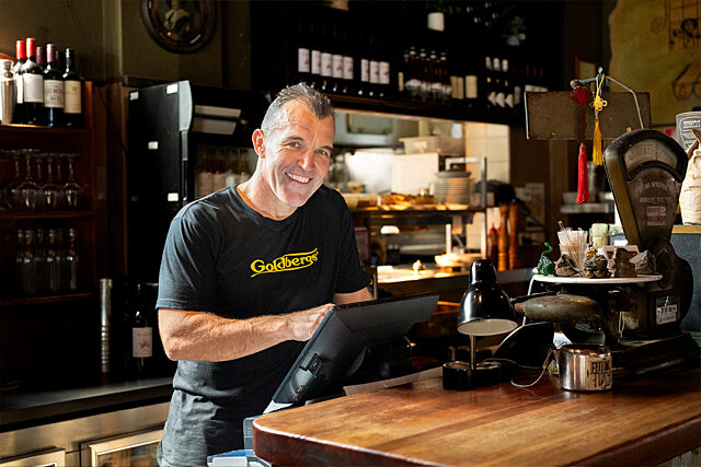 Person in a 'Goldberg's' t-shirt using a touchscreen at a restaurant counter; shelves with bottles, glasses, and kitchen equipment in the background.
