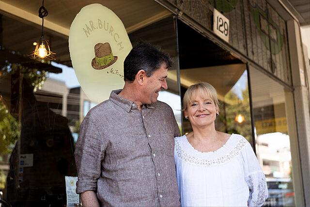 Two people standing in front of a storefront with a 'MR. BURCH' sign featuring a hat illustration and 'est. 1979'; address number '129' above the entrance.
