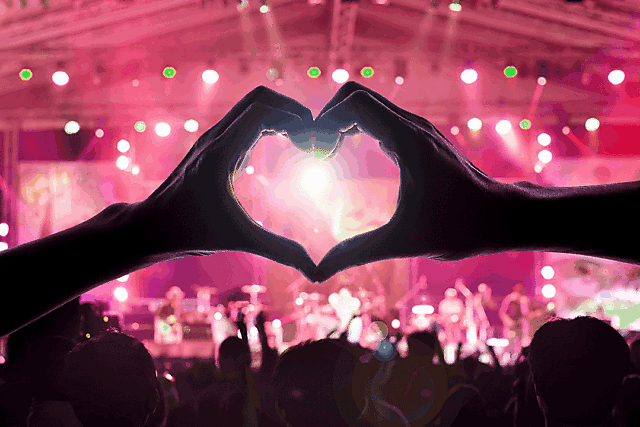 Hands forming a heart shape with fingers, framing a brightly lit concert stage.