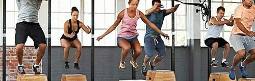 Five people mid-air performing box jumps onto wooden boxes in a gym with large windows and equipment.