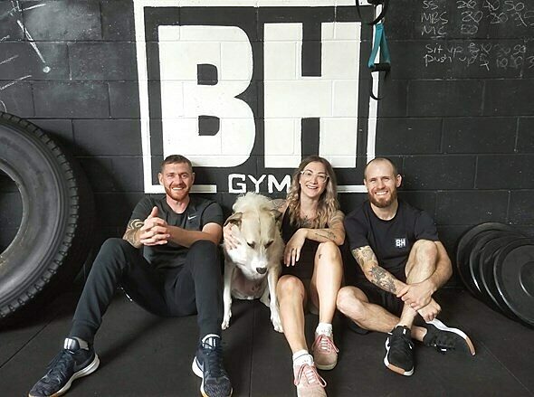 Three people and a dog seated in front of a “BH GYM” sign at Brickhouse Gym, with large tires and weight plates nearby.