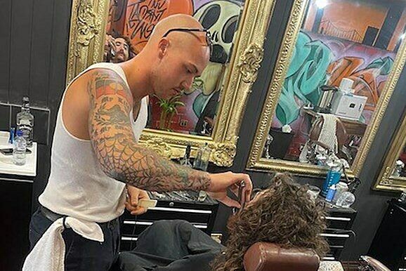 Tattooed barber in a white tank top styling a client's hair in a salon with ornate mirrors and hairdressing tools.