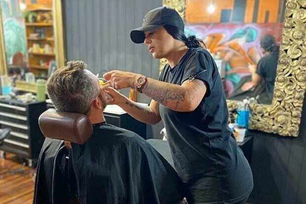 Person seated in a barber chair with a black cape while a barber works on their hair in a salon setting.