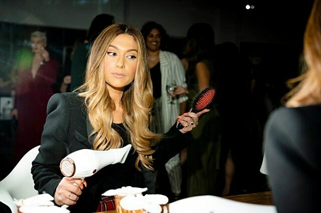 Natalie Anne holding a hairdryer and brush in a dimly lit room with people in the background.