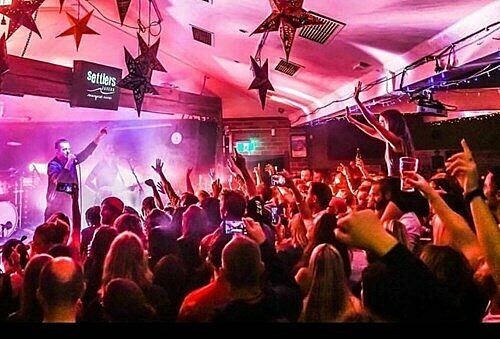 Performer on stage at Settlers Tavern with microphone, colourful lights, and crowd raising hands under star decorations.