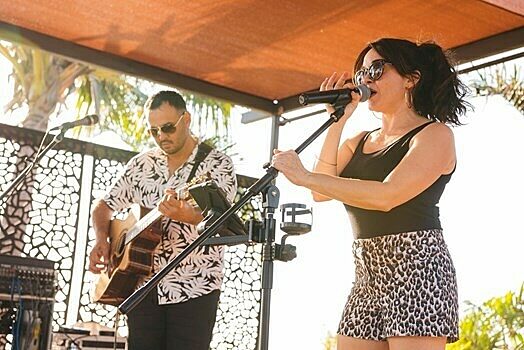 Acoustic guitarist and singer performing on outdoor stage at Darwin Ski Club with palm trees in background.