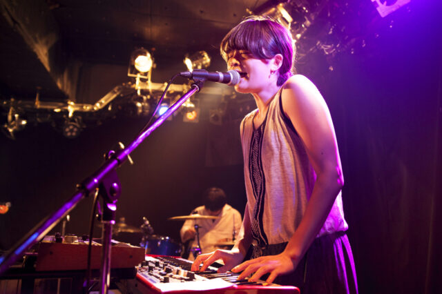 Musician playing keyboard and singing on stage with drummer in background under vibrant lighting.