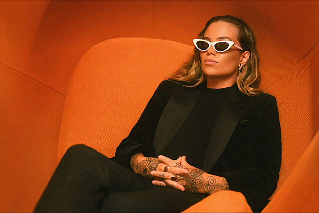 Australian songwriter and performer Tash Sultana, sitting in a bright orange chair wearing all black with white sunglasses.