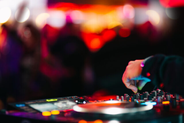 DJ adjusting mixing console with colourful lights and crowd in background.