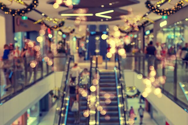 Blurred view of a shopping mall with escalators, lights, garlands, and people on both levels.