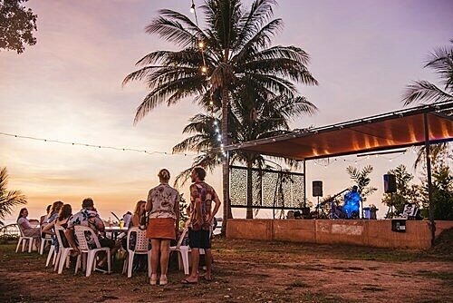 Group of people gathered outdoors near a stage with musical equipment, under palm trees and string lights at sunset.