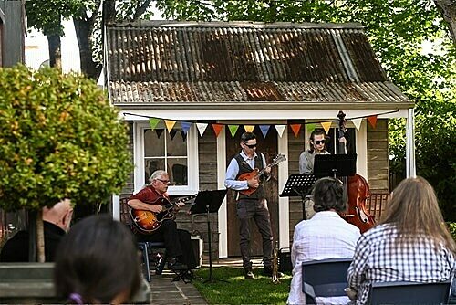 Outdoor musical performance with four musicians in front of a shed, watched by seated audience.