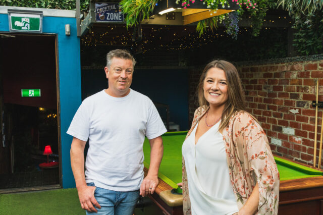 Two people stand beside a pool table in an indoor space with a brick wall, greenery, and a “FOOTBALL ZONE” sign in the background. An emergency exit sign is visible on the left.
