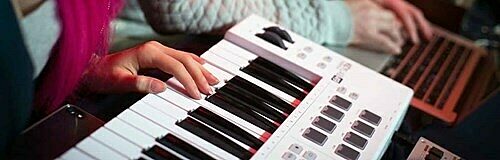 Close-up of hand playing white and black keyboard with person using laptop in background.