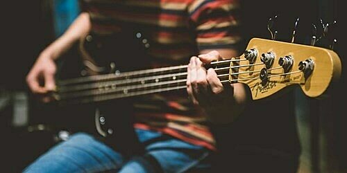 Person playing electric bass guitar, wearing striped shirt and jeans, with focus on hands and headstock.