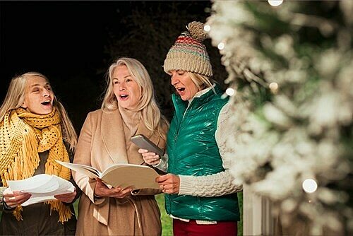 Three people in winter clothing singing carols from song sheets beside a decorated Christmas tree.