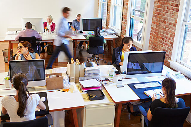 Busy office with people working at desks, exposed brick walls, and large windows.